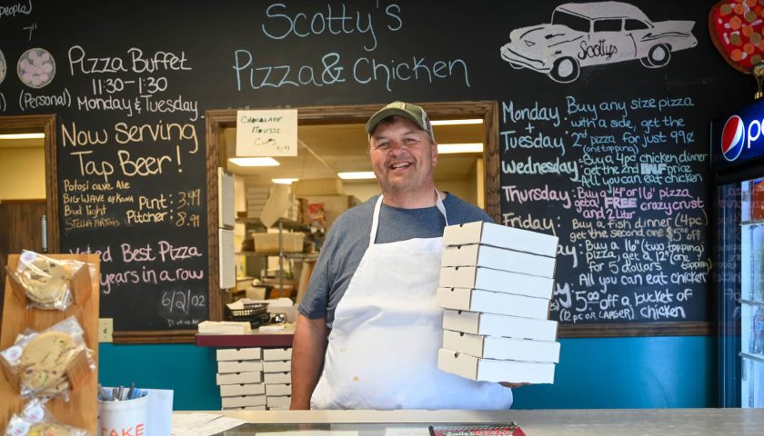 Find comfort food & cozy lodging in Marshfield this winter | Owner at Scotty’s Pizza & Chicken in Marshfield Wisconsin holding pizza boxes at counter